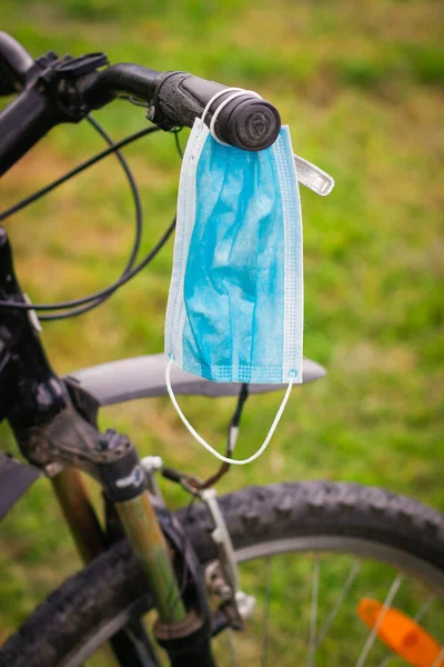 A blue medical Mask hangs on handlebar of Bicycle against background of green Grass. Sports and walking during self-isolation.