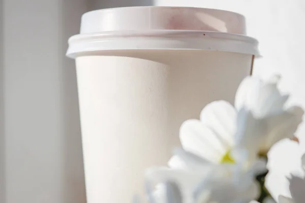 Coffee to go with flowers. Take away cup with shabby flowers on white sunny background. Coffee or tea paper cup.