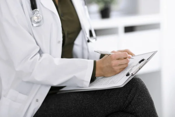 Mujer médica desconocida escribiendo algo en el portapapeles mientras está sentada en la silla, de cerca. El terapeuta que usa blusa verde en el trabajo está llenando el historial de medicamentos. Concepto de medicina — Foto de Stock