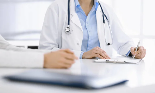 Okänd kvinna-läkare och kvinnlig patient sitter och talar vid läkarundersökning på kliniken, närbild. Terapeut klädd i blå blus fyller upp medicinhistoria rekord. Medicinskt koncept — Stockfoto