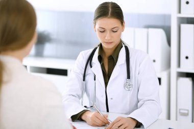 Kadın doktor ve hasta hastane muayenehanesinde oturup konuşuyorlar. Yeşil renkli bluz terapiste yakışır. Tıp ve sağlık hizmetleri kavramı