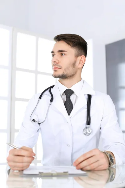 Medico che compila la scheda della storia dei farmaci mentre è seduto alla scrivania di vetro nello studio medico o in clinica. Medicina e concetto di assistenza sanitaria — Foto Stock