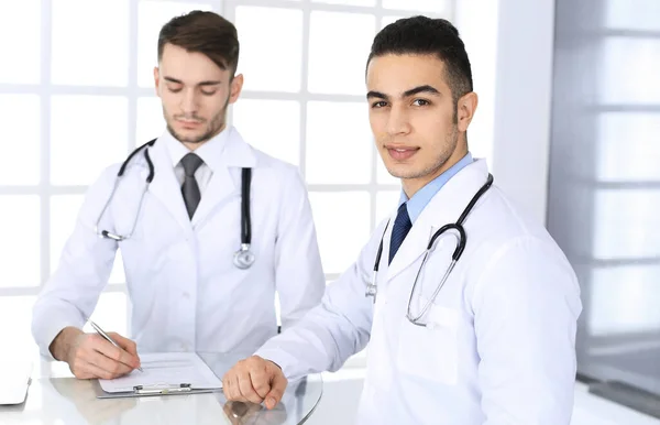 Arabische arts die aan het glazen bureau zit met een blanke collega in een medisch kantoor of kliniek. Verscheidene artsen team, geneeskunde en gezondheidszorg concept — Stockfoto