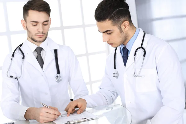 Arabski lekarz siedzący przy szklanym biurku z białym kolegą w gabinecie medycznym lub klinice. Różnorodny zespół lekarzy, koncepcja medycyny i opieki zdrowotnej — Zdjęcie stockowe