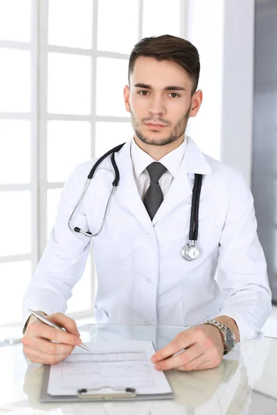 Médecin homme remplir les dossiers de l'histoire des médicaments formulaire tout en étant assis au bureau de verre dans le bureau médical ou clinique. Médecine et concept de santé — Photo