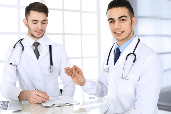 Arabische arts die aan het glazen bureau zit met een blanke collega in een medisch kantoor of kliniek. Verscheidene artsen team, geneeskunde en gezondheidszorg concept — Stockfoto