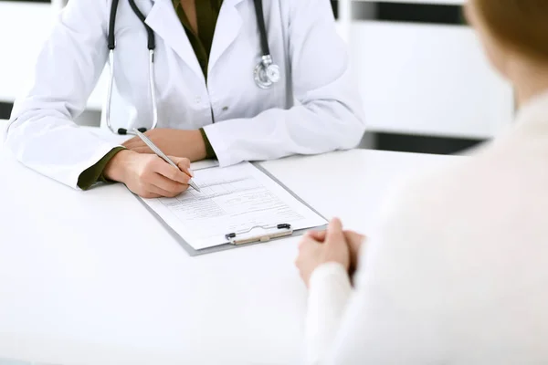 Vrouwelijke dokter en patiënt zitten en praten bij medisch onderzoek op kantoor, close-up. Therapeut vult medicijngeschiedenis dossiers bij. Concept geneeskunde en gezondheidszorg — Stockfoto