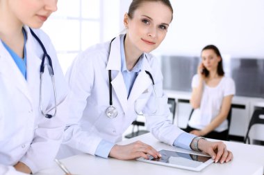 Hastanede çalışan bir grup doktor ve arka planda sırada bekleyen bir hasta. Doktor resepsiyonda beklerken tıbbi belge ya da reçete dolduruyor. Tıp ve sağlık verileri