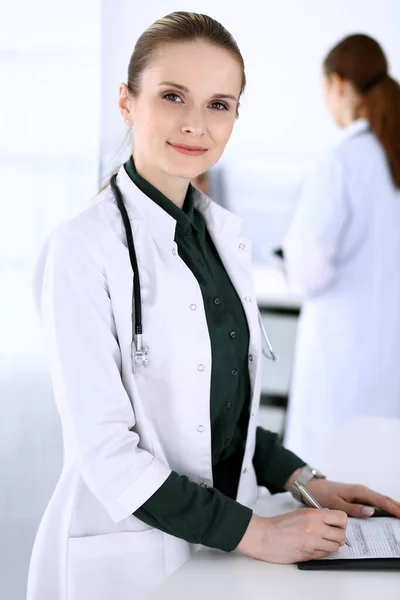 Doctora mujer en el trabajo con el paciente y colega en el fondo. Médico llenando documentos médicos o prescripción mientras está de pie en la recepción del hospital. Datos en medicina y asistencia sanitaria — Foto de Stock