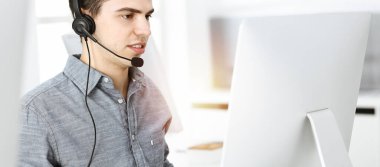 Güneşli ofiste müşterilerle konuşurken kulaklık ve bilgisayar kullanan sıradan giyinmiş genç bir adam. Çağrı merkezi, iş konsepti