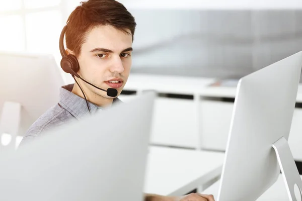 Hombre joven vestido casual usando auriculares y computadora mientras habla con los clientes en línea en la oficina soleada. Call center, concepto de negocio —  Fotos de Stock