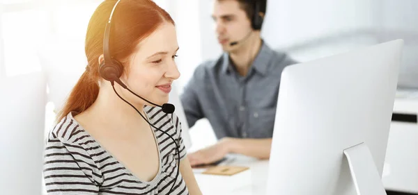 在网上与客户交谈时，用耳机和电脑随意穿衣的年轻女性。一群操作员在阳光灿烂的办公室里工作.呼叫中心 — 图库照片
