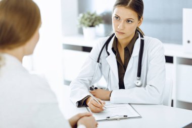 Kadın doktor ve hasta hastane ofisinde oturuyor ve konuşuyor. Yeşil renkli bluz terapiste uygun.