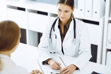 Kadın doktor ve hasta hastane ofisinde oturuyor ve konuşuyor. Yeşil renkli bluz terapiste uygun.