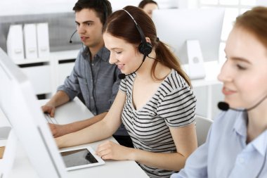 İnternette müşterilerle konuşurken kulaklık ve bilgisayar kullanan sıradan giyinmiş genç bir kadın. Bir grup operatör iş başında. Çağrı merkezi, iş konsepti