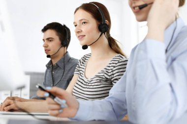 İnternette müşterilerle konuşurken kulaklık ve bilgisayar kullanan sıradan giyinmiş genç bir kadın. Bir grup operatör iş başında. Çağrı merkezi, iş konsepti