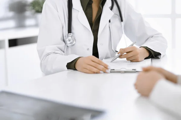 Onbekende vrouw-dokter en vrouwelijke patiënt zitten en praten bij medisch onderzoek in de kliniek, close-up. Therapeut met groene blouse vult de geschiedenis van medicatie. Geneesmiddelenconcept — Stockfoto