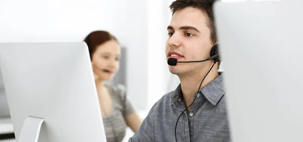 Casual vestido jovem usando fone de ouvido e computador enquanto conversa com clientes on-line. Grupo de operadores a trabalhar. Centro de atendimento — Fotografia de Stock