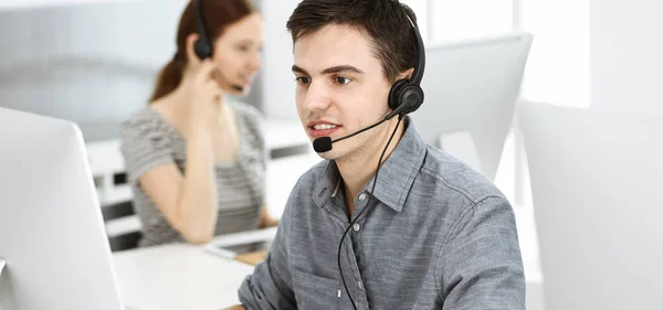 Hombre joven vestido casual usando auriculares y computadora mientras habla con los clientes en línea. Grupo de operadores en el trabajo. Centro de llamadas —  Fotos de Stock