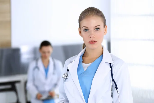 Doctor woman standing in emergency hospital office with colleague at background. Physician at work, studio portrait. Medicine and health care concept
