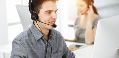 Güneşli ofiste müşterilerle konuşurken kulaklık ve bilgisayar kullanan sıradan giyinmiş genç bir adam. Çağrı merkezi, iş konsepti