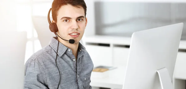 Nenuceně oblečený mladý muž pomocí sluchátka a počítače při rozhovoru se zákazníky on-line ve slunné kanceláři. Call centrum, obchodní koncept — Stock fotografie