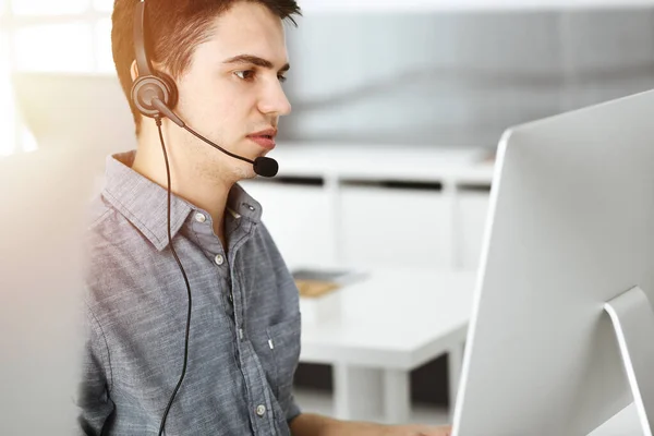 Casual vestido jovem usando fone de ouvido e computador enquanto conversa com clientes on-line no escritório ensolarado. Call center, conceito de negócio — Fotografia de Stock