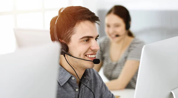 Hombre joven vestido casual usando auriculares y computadora mientras habla con los clientes en línea en la oficina soleada. Call center, concepto de negocio —  Fotos de Stock