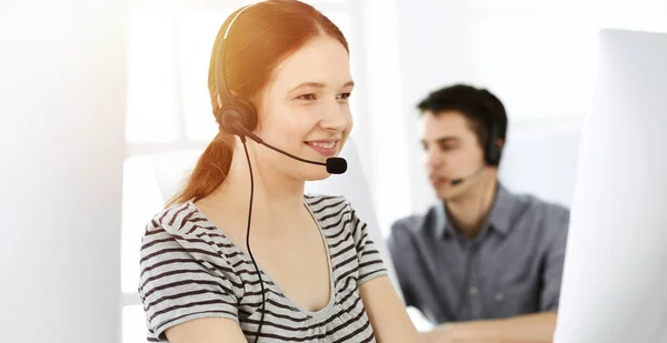 Casual dressed young woman using headset and computer while talking with customers online. Group of operators at work in sunny office. Call center