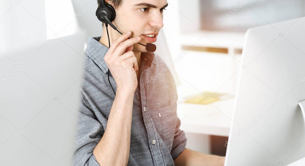 Casual dressed young man using headset and computer while talking with customers online in sunny office. Call center, business concept