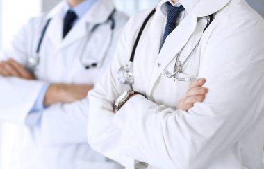 Bir grup modern doktor hastane ofisinde kollarını kavuşturmuş bir takım gibi yakın plan çekimde duruyorlar. Doktorlar hastaları muayene etmeye ve yardım etmeye hazır. Tıbbi yardım, sağlık sigortası, en iyi tedavi ve