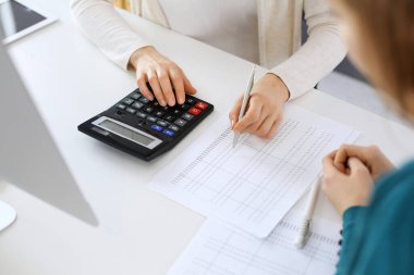 Muhasebeci mali durumu kontrol ediyor ya da vergi formları için hesap makinesi geliri ile sayıyor, eller yakın çekim. İş kadını ofisteki masada iş arkadaşıyla oturuyor ve çalışıyor. Denetim kavramı