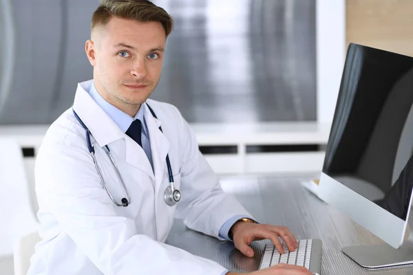 Doctor hombre sentado detrás de una computadora en el lugar de trabajo en la clínica u oficina del hospital. Fotografía médica y concepto de medicina — Foto de Stock