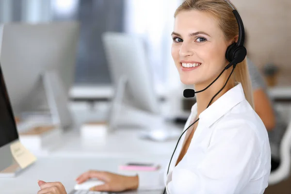 顧客サービスオフィスでのコミュニケーションやコンサルティングにヘッドセットを使用してブロンドのビジネス女性。コールセンター。バックグラウンドでの作業者のグループ — ストック写真