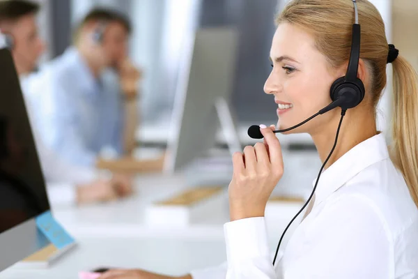 Mujer rubia de negocios con auriculares para la comunicación y la consulta de las personas en la oficina de servicio al cliente. Centro de llamadas. Grupo de operadores en el trabajo en segundo plano — Foto de Stock