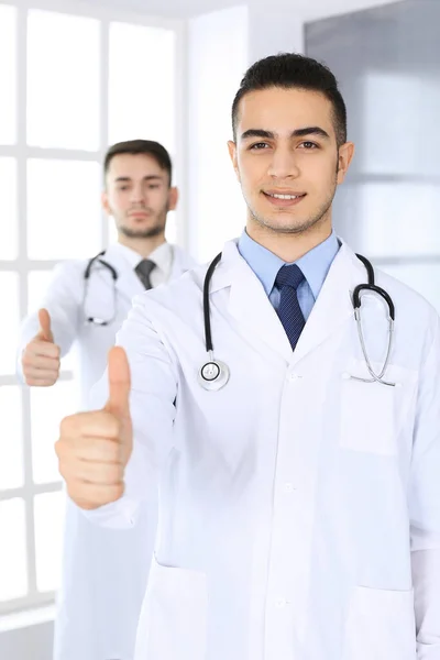 Homem médico árabe mostrando Ok assinar com polegares para cima com colega caucasiano em consultório médico ou clínica. Equipe de médicos diversos, melhor tratamento, medicina e conceito de saúde — Fotografia de Stock