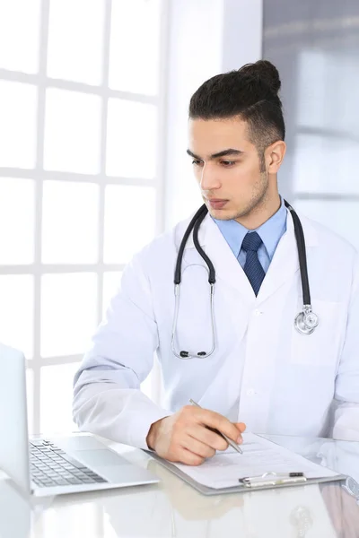 Арабский врач, пользуясь ноутбуком, заполняет анкету в стеклянном столе в медицинском кабинете или клинике. Медицина и здравоохранение — стоковое фото
