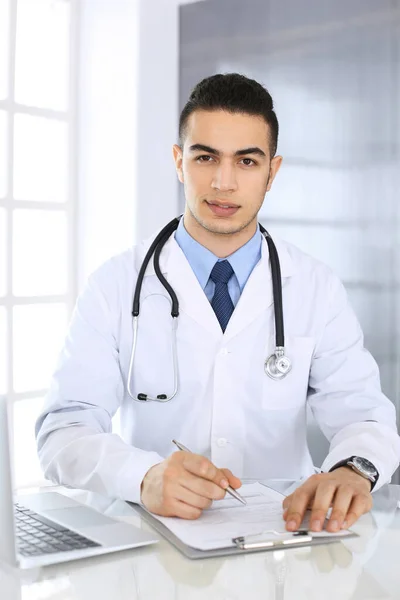 Hombre médico árabe que usa computadora portátil mientras llena el formulario de registros de historial de medicamentos en el mostrador de vidrio en la oficina médica o clínica. Concepto de medicina y salud —  Fotos de Stock
