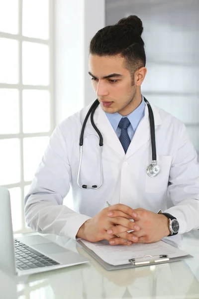 Arabe médecin homme utilisant un ordinateur portable tout en remplissant les dossiers d'historique de médicaments formulaire au bureau de verre dans le bureau médical ou clinique. Médecine et concept de santé — Photo