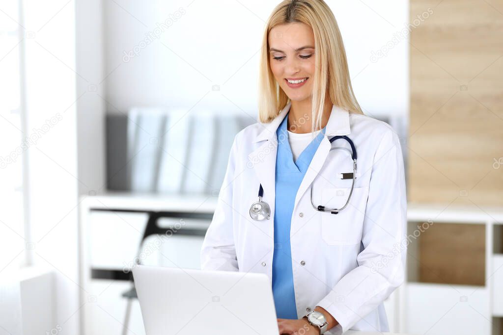 Doctor woman at work in hospital excited and happy of her profession. Blonde physician controls medication history records and exam results while using laptop computer. Medicine and healthcare concept