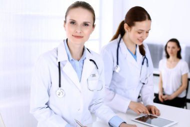 Hastanede çalışan bir grup doktor ve arka planda sırada bekleyen bir hasta. Doktor resepsiyonda beklerken tıbbi belge ya da reçete dolduruyor. Tıp ve sağlık verileri