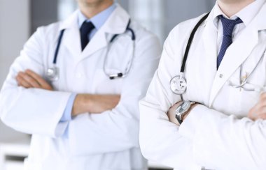Bir grup modern doktor hastane ofisinde kollarını kavuşturmuş bir takım gibi yakın plan çekimde duruyorlar. Doktorlar hastaları muayene etmeye ve yardım etmeye hazır. Tıbbi yardım, sağlık sigortası, en iyi tedavi ve