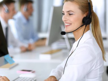 Sarışın iş kadını iletişim kurmak ve müşteri hizmetlerindeki insanlara danışmak için kulaklık kullanıyor. Merkezi ara. Arka planda çalışan bir grup operatör