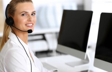 Merkezi ara. İnternette müşterilere danışırken kulaklık kullanan mutlu ve heyecanlı bir iş kadını. Müşteri hizmetleri ofisi ya da tele pazarlama bölümü. İşte gülümseyen sarışın kadın operatör