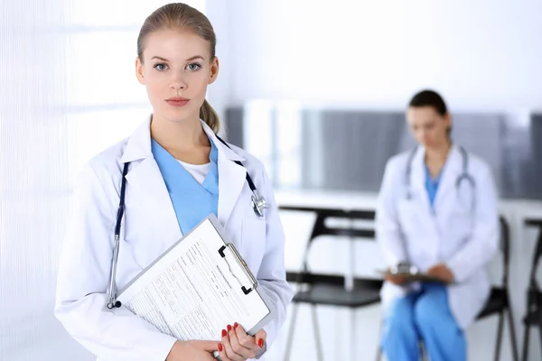 Doctor woman standing in emergency hospital office with colleague at background. Physician at work, studio portrait. Medicine and health care concept