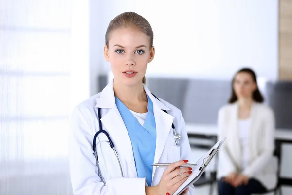 Orvos nő kitölti a kórtörténetét, miközben a sürgősségin áll egy beteggel a háttérben. Orvos a munkahelyén, portré fotózás. Orvosi és egészségügyi ellátás fogalma — Stock Fotó