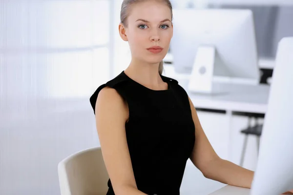 Business woman working with computer while sitting at the desk in modern office. Secretary or female lawyer looks beautiful in black dress. Business people concept