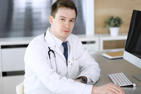 Dokter man zit achter een computer op het werk in kliniek of ziekenhuis kantoor. Medisch hoofdschot en medicijnconcept — Stockfoto