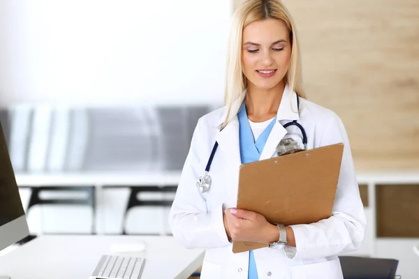 Doctora mujer en el trabajo en el hospital emocionada y feliz de su profesión. Médico rubio controla los registros de la historia de la medicación y los resultados del examen mientras usa portapapeles. Concepto de medicina y salud — Foto de Stock