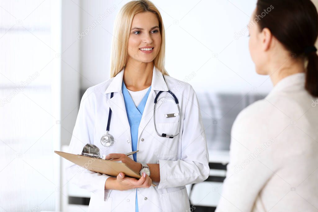 Doctor woman at work in hospital excited and happy of consulting female patient. Blonde physician controls medication history records and exam results while using clipboard. Medicine and healthcare